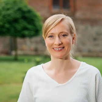 Eine Frau mit einem hellen Pullover mit V-Ausschnitt lächelt in die Kamera. Sie hat blonde Haare, nur ihr Oberkörper ist zu sehen. Im Hintergrund etwas verschwommen ist eine Rasenfläche und der untere Teil eines Gebäudes aus Ziegelstein zu sehen.